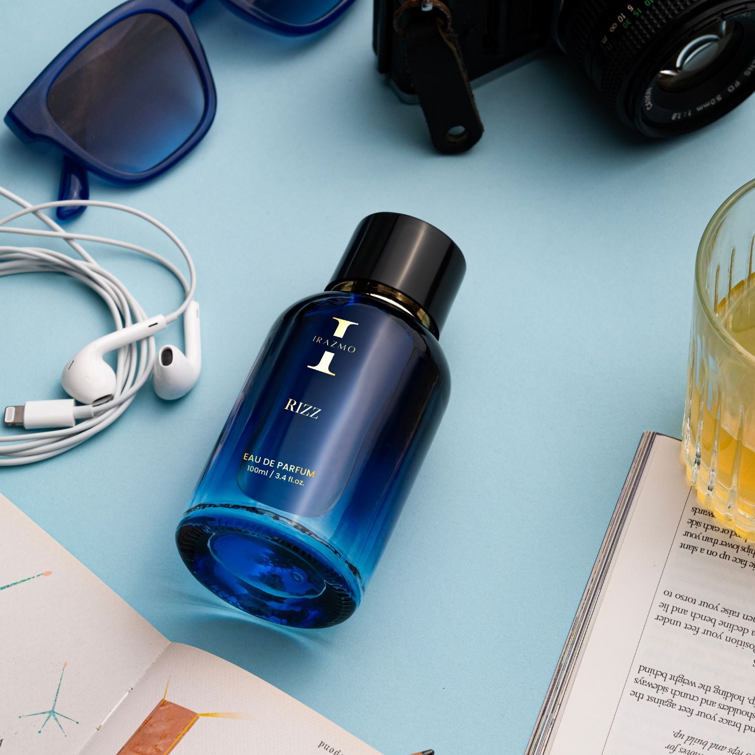 Blue perfume bottle on a light blue surface with sunglasses, camera, earphones, and a glass of orange juice.