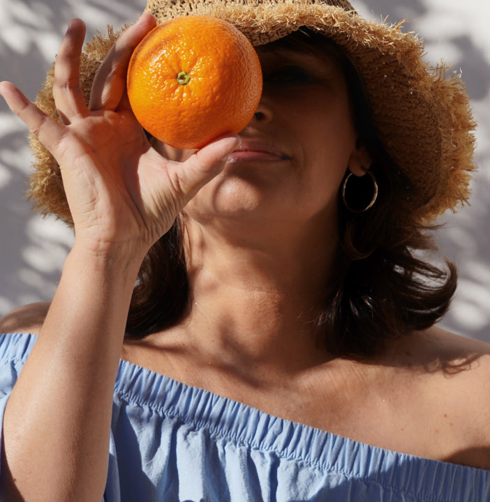 Woman holding an orange in front of her face with a straw hat and blue top.