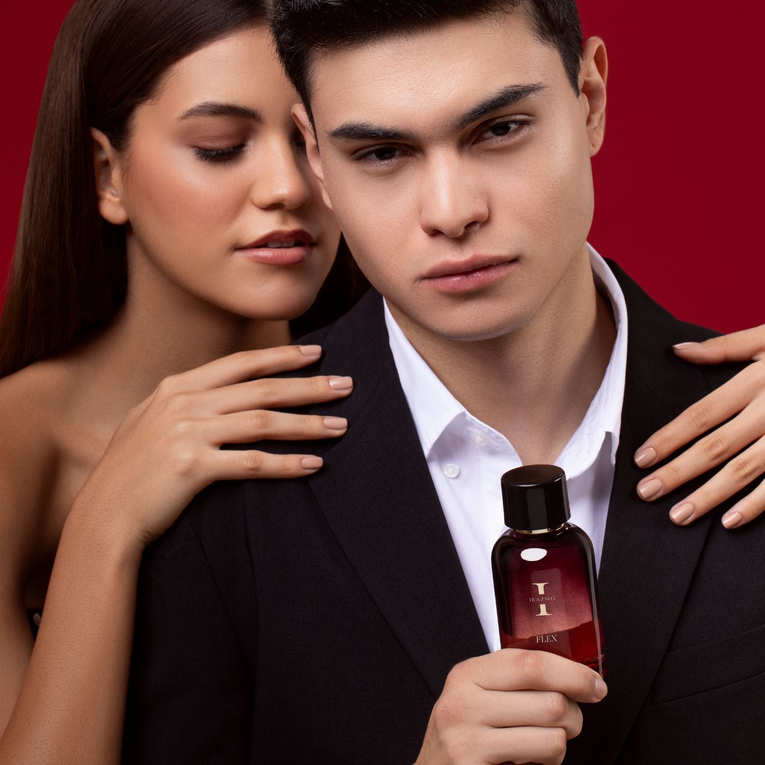 Man holding a perfume bottle with a woman's hand on his shoulder against a red background