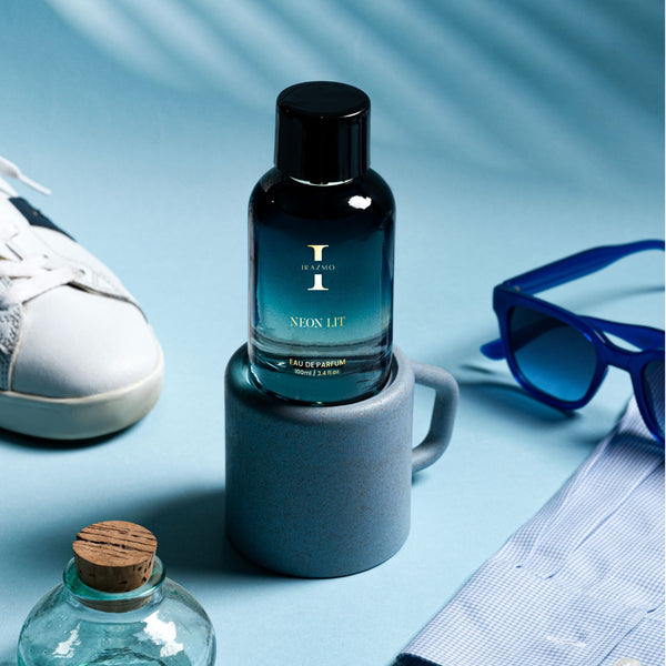 Perfume bottle labeled 'Neon Lit' on a blue surface with a shoe, sunglasses, and other items.